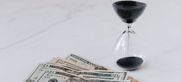 An hourglass with black sand next to US dollar bills.