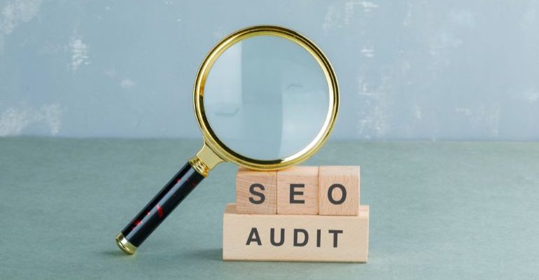 Magnifying glass leaning on SEO AUDIT spelled out with wooden cubes.