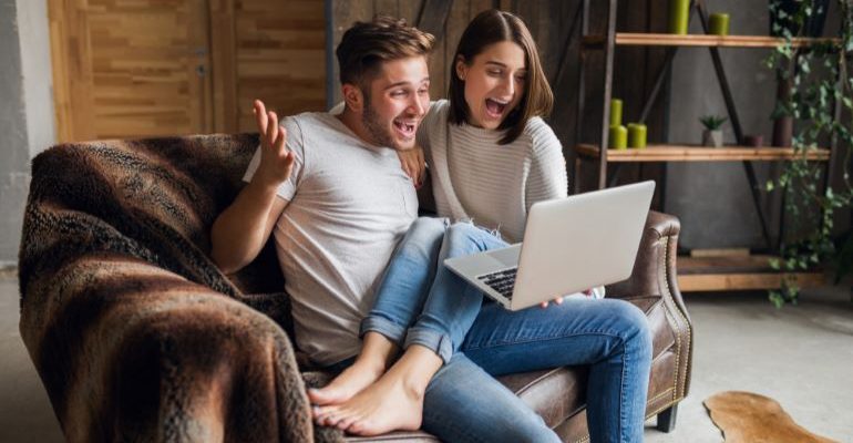 A couple sitting on a couch and looking at a laptop, thrilled.