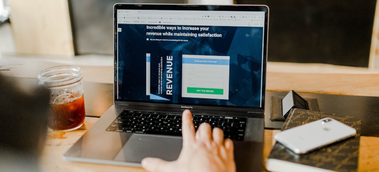Person pointing a finger at a laptop monitor located on a desk between a glass and a a smartphone 