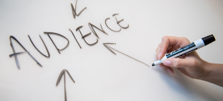 Person using a black marker to draw arrows pointing towards the word audience written on a white board
