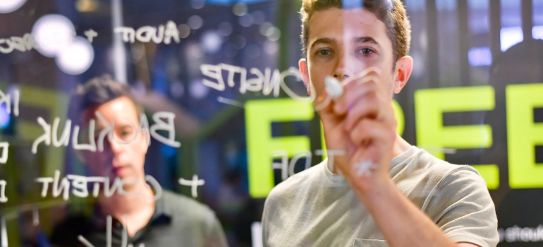 Two people using a white marker to write on a glass wall