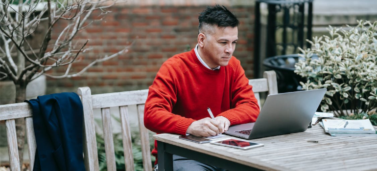 A man in a red shirt working on a laptop outdoors.