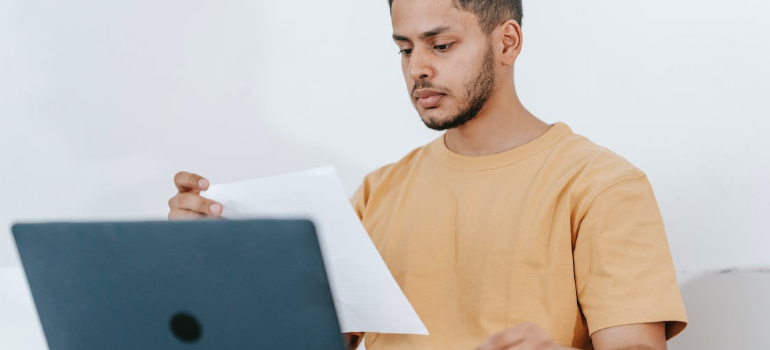 A man looking at a piece of paper in front of a laptop.