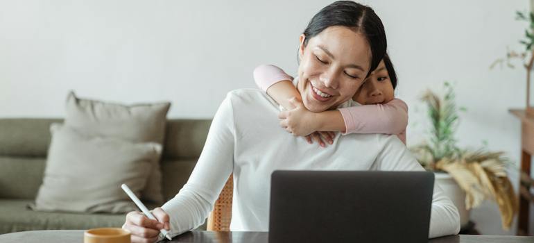 A child hugging her mother while she is using a laptop