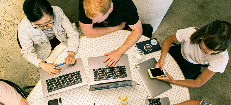 A few people sitting on a round desk using their laptops and phones.