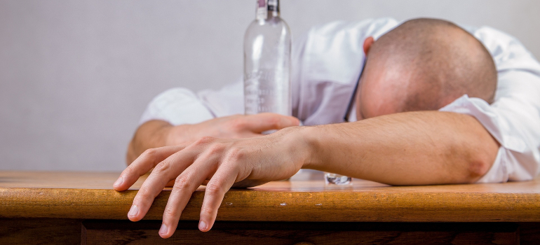 A man lying on a desk with an empty alcohol bottle next to him. 