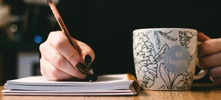 Hand holding a pen and writing, mug in the other hand