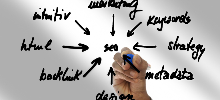 A hand writing on a transparent surface, showing a diagram with "SEO" at the center and arrows pointing to related terms like "marketing," "keywords," "strategy," "metadata," "design," "backlink," "HTML," and "content."