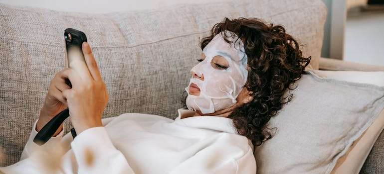 A person lying on a couch wearing a sheet mask and using a smartphone